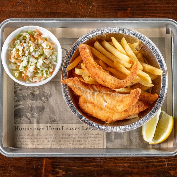 Fried fish and french fries in a foil container with coleslaw and lemon wedges on a tray.