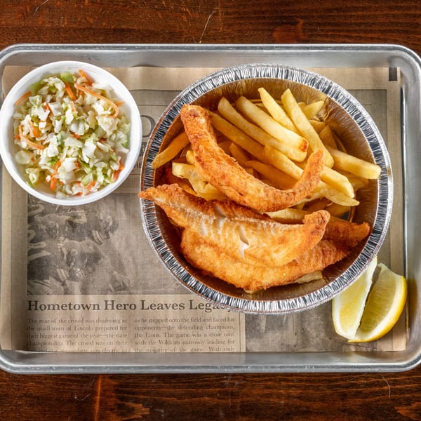 Fried fish and french fries in a foil container with coleslaw and lemon wedges.
