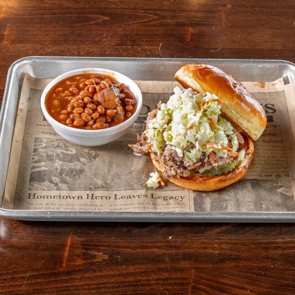 Pulled pork sandwich with coleslaw and pickles, served with a side of baked beans on a tray.